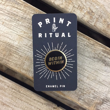 Black circle enamel pin with "begin within" in gold with gold border on print ritual card backing