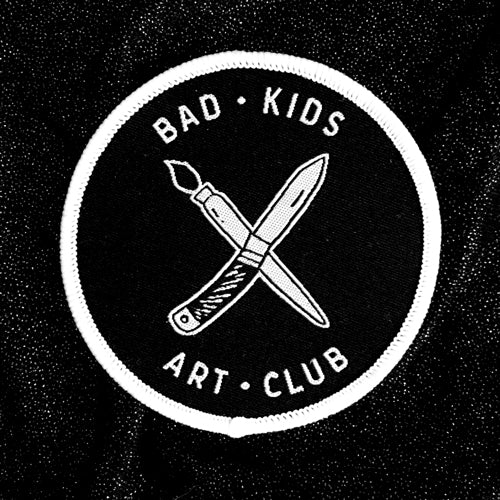 Circle iron on patch with &quot;x&quot; of paintbrush and knife with Bad Kids Art Club around it embroidered in white. Merrowed edge in white. By Print Ritual